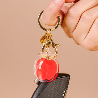 A hand holds The Darling Effect’s Apple Acrylic Keychain—a cute, durable accessory featuring a shiny red apple charm and gold bell—attached to a car key and standing out against a neutral beige background.