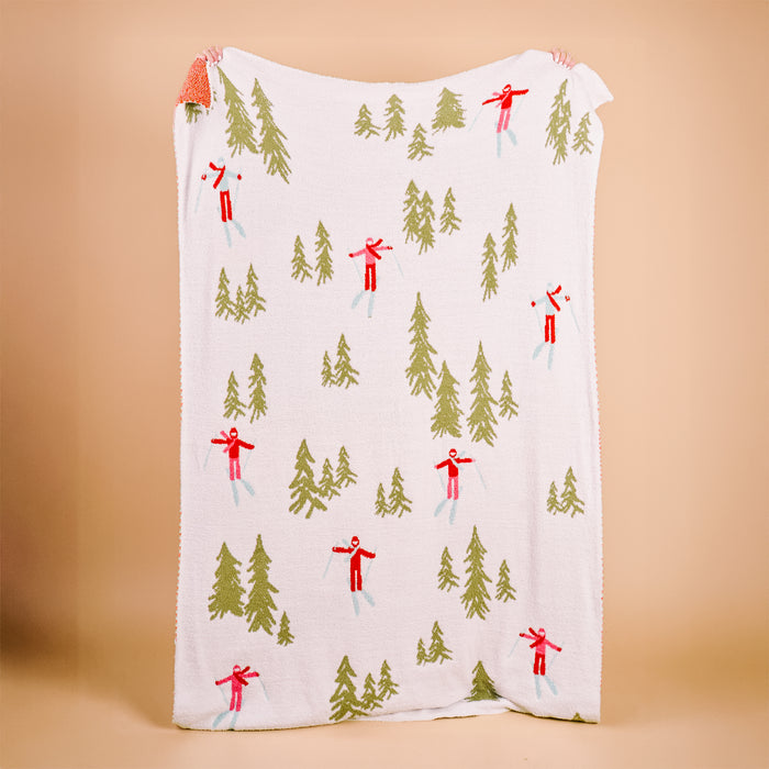 The Darling Effect Aprés Ski Cozy Dreams Luxe Blanket, featuring green trees and red skiers on white, is held by two hands at each top corner against a plain beige background.