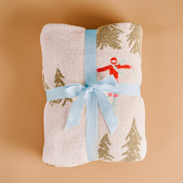 The Darling Effect Aprés Ski Cozy Dreams Luxe Blanket features a fuzzy white, ultra-soft polyester microfiber with a winter print of snow-covered trees and a skier, finished with a pale blue ribbon.