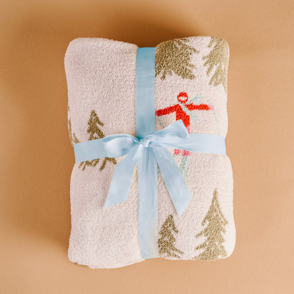 The Darling Effect Aprés Ski Cozy Dreams Luxe Blanket features a fuzzy white, ultra-soft polyester microfiber with a winter print of snow-covered trees and a skier, finished with a pale blue ribbon.