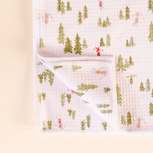 The Darling Effect's Aprés Ski Waffle Tea Towel features a white cotton waffle design with small green trees and skiers in red and blue outfits, shown with one corner folded to display the underside.