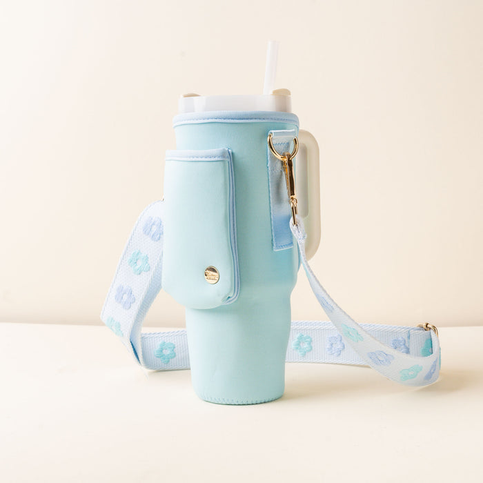 A light blue tumbler sling with an adjustable strap and pocket, holding a white tumbler with a straw. The hands-free strap showcases blue and teal flowers. The background is off-white.