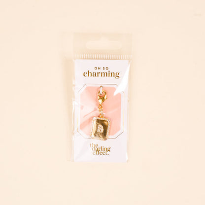The-darling-effect B Jewelry Charm Initial Letter Gold features a gold-plated "B" charm on a white and peach card with "oh so charming" and "the darling effect," packaged in a clear plastic bag.