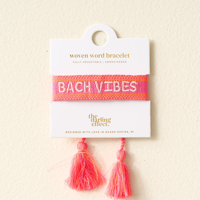 The Darling Effect's Bach Vibes Woven Bracelet features pink adjustable cords, colorful embroidery spelling "BACH VIBES" in white, and two pink tassels. It comes displayed on a white card with gold text for a stylish presentation.