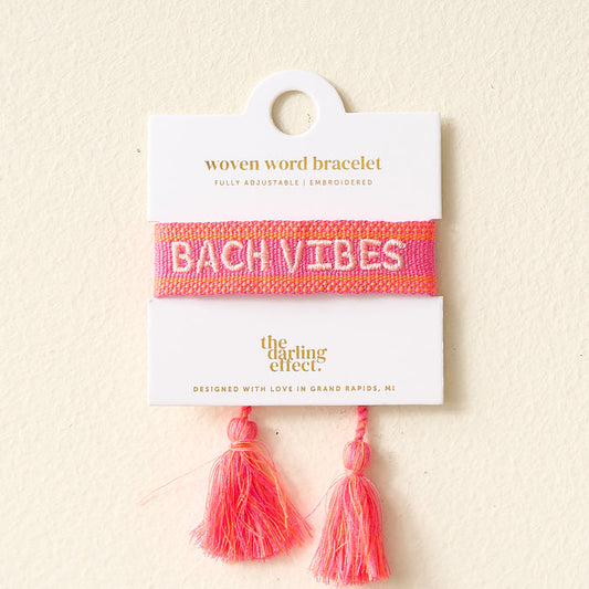 The Darling Effect's Bach Vibes Woven Bracelet features pink adjustable cords, colorful embroidery spelling "BACH VIBES" in white, and two pink tassels. It comes displayed on a white card with gold text for a stylish presentation.