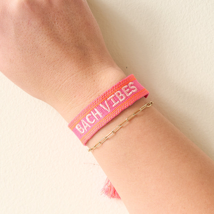 A hand sports The Darling Effect's Bach Vibes Woven Bracelet—pink with white text—stacked next to a delicate gold chain bracelet, perfect for creating a trendy wrist look.