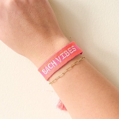 A hand sports The Darling Effect's Bach Vibes Woven Bracelet—pink with white text—stacked next to a delicate gold chain bracelet, perfect for creating a trendy wrist look.