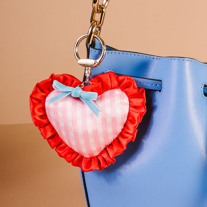 A blue handbag with The Darling Effect’s Berry Sweet Puffy Heart Bag Charm—a pink heart-shaped accessory with red ruffles and a blue bow—attached to the gold clasp, set against a soft beige background.
