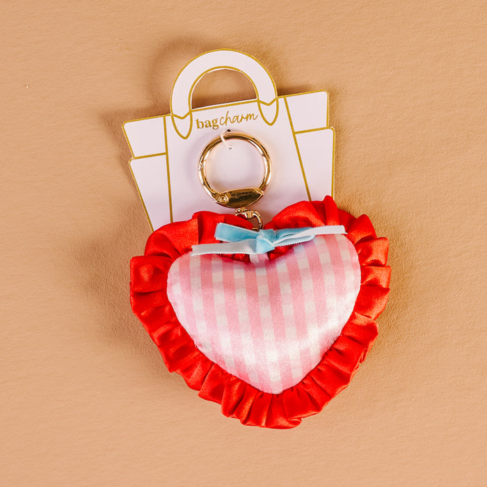 The Darling Effect's Berry Sweet Puffy Heart Bag Charm features a gold keyring, red ruffled edges, pink and white stripes, and a blue bow—making it a cute accessory for your bag. Shown against a beige background.