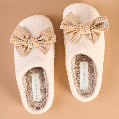 The Darling Effect's Betty Bow Maple Gingham Fuzzy Slippers feature a fluffy cream exterior, soft gray lining, and large beige and white gingham bows, displayed on a light brown surface.