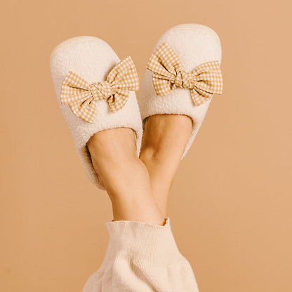 A person with crossed legs in light pants wears The Darling Effect's Betty Bow Maple Gingham Fuzzy Slippers, featuring cozy fall vibes and large checkered bows, set against a beige background.