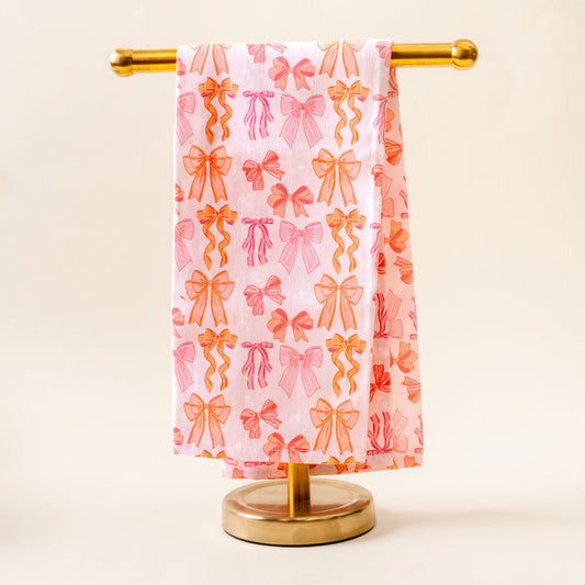 The Blushing Bows Tea Towel by the-darling-effect, featuring pink, orange, and red bows on white cotton, hangs neatly on a gold stand against a simple light background.