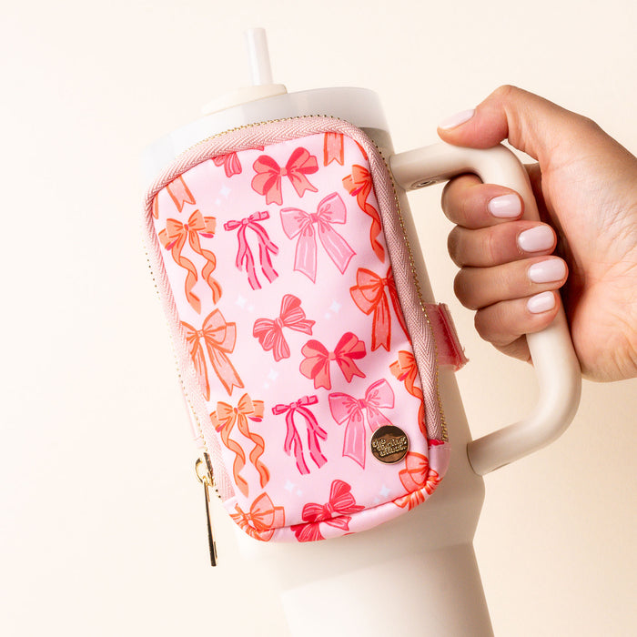 A hand holds a white tumbler with a handle, featuring the-darling-effect’s Blushing Bows Tumbler Fanny Pack—a water-resistant pink pouch with red and orange bow designs. The person sports neatly manicured, pale nails.
