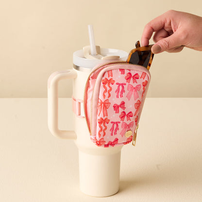 A hand places sunglasses into the Blushing Bows Tumbler Fanny Pack by the-darling-effect, a stylish pink, water-resistant pouch with a bow pattern that fits on a large cream tumbler with straw and handle.