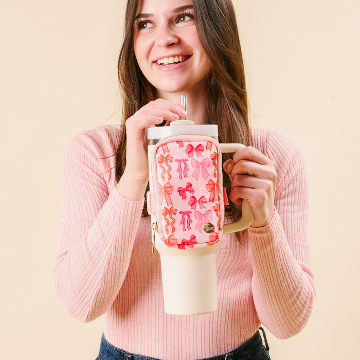 A woman in a pink sweater holds a large cream tumbler with the-darling-effect’s Blushing Bows Tumbler Fanny Pack attached. She looks slightly off camera, smiling, and has a metal straw in hand.