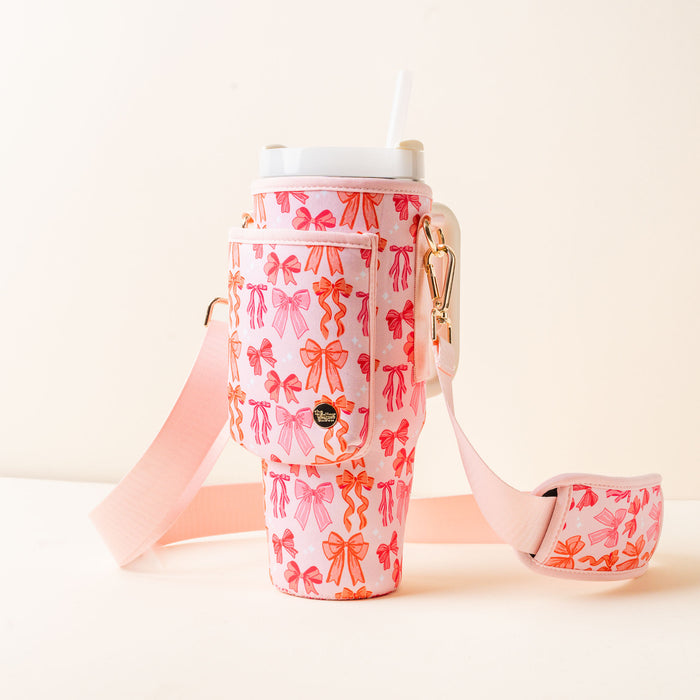 The-darling-effect’s Blushing Bows Tumbler Sling features a pink and orange bow pattern, pocket, gold hardware, hands-free adjustable pink strap, a white straw, and is perfect for stylish sipping on the go.