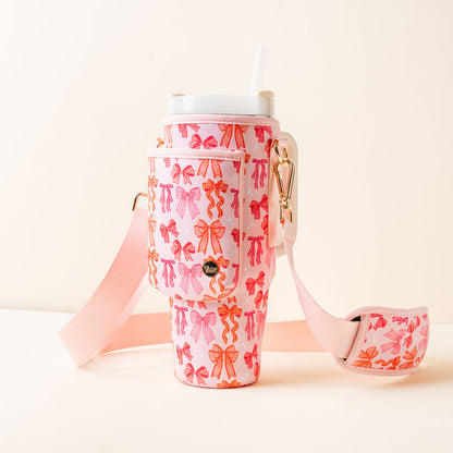 The-darling-effect’s Blushing Bows Tumbler Sling features a pink and orange bow pattern, pocket, gold hardware, hands-free adjustable pink strap, a white straw, and is perfect for stylish sipping on the go.