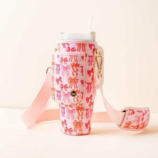The-darling-effect’s Blushing Bows Tumbler Sling features a pink and orange bow pattern, pocket, gold hardware, hands-free adjustable pink strap, a white straw, and is perfect for stylish sipping on the go.
