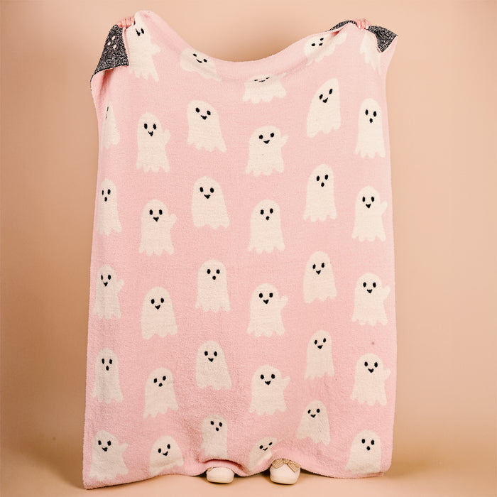 An unseen person holds up The Darling Effect's Boo Crew Cozy Dreams Luxe Blanket, a pink ultra-soft microfiber throw with white cartoon ghosts and a stylish black and white patterned edge.