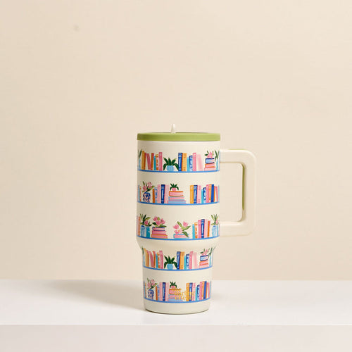 The Darling Effect’s Book Shelfie 20 oz Lifestyle Flip Straw Tumbler, featuring a cream finish, large handle, and colorful book and plant illustrations on three shelf rows, rests on a white surface with a light beige background.
