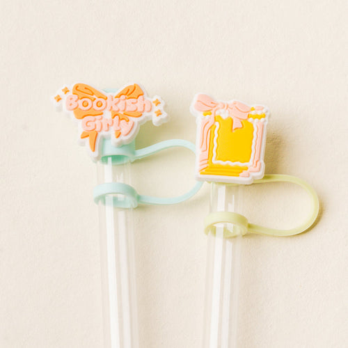 The Bookish Girly Straw Topper Set by the-darling-effect features two clear straws with pastel silicone toppers: an orange bow with "Bookish Girly" text and a yellow present with pink bow—fun drinkware accessories for any occasion.