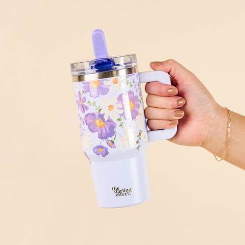A hand with manicured nails holds a Bouquet Beauty Purple 20 oz Lifestyle Flip Straw Tumbler from the-darling-effect, featuring a lavender and white design, cream background, lid, purple straw, and logo on the front.