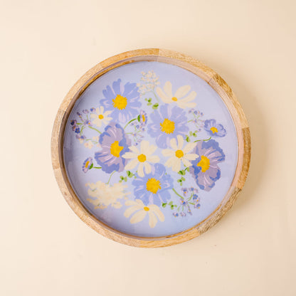 The-darling-effect’s Bouquet Beauty Purple Large Round Wood Enamel Tray features a painted floral design in blue, white, and yellow on a light purple background, displayed against a cream-colored wall.