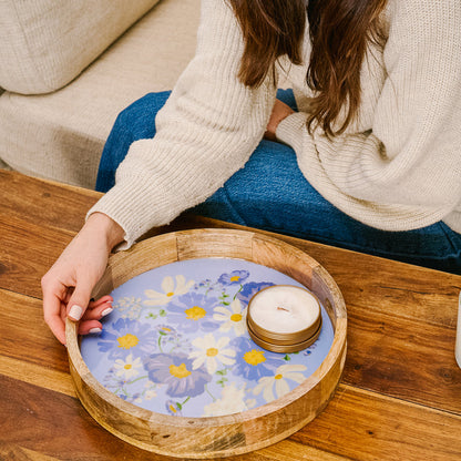 A person in a cream sweater and blue jeans places the-darling-effect's Bouquet Beauty Purple Large Round Wood Enamel Tray with a lit candle on a wooden table.