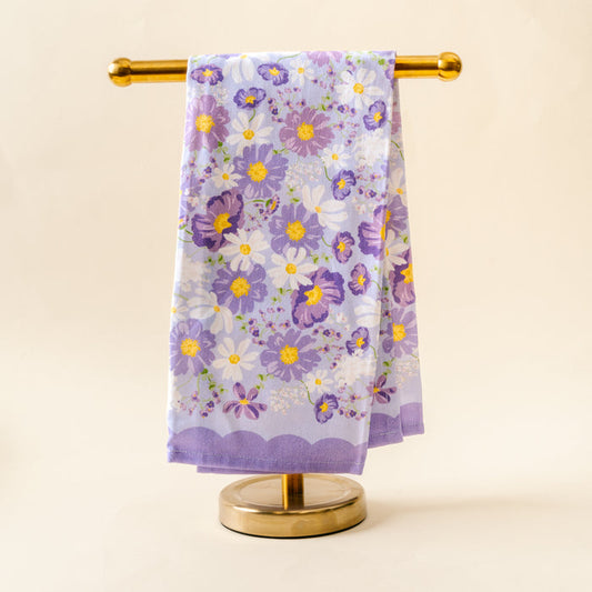 The Bouquet Beauty Purple Tea Towel by the-darling-effect, featuring a vibrant floral pattern in purple, yellow, and white cotton, hangs neatly on a gold towel stand against a light beige background.