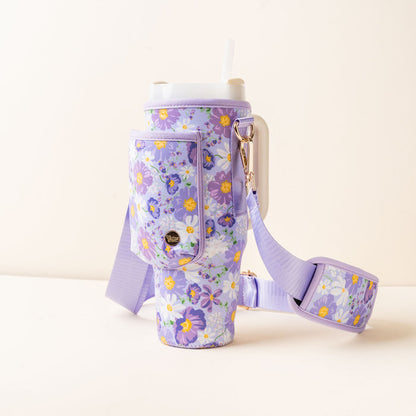 A large tumbler with a straw is in the-darling-effect's Bouquet Beauty Purple Tumbler Sling, which features a pocket and an adjustable hands-free strap, displayed against a plain light background.