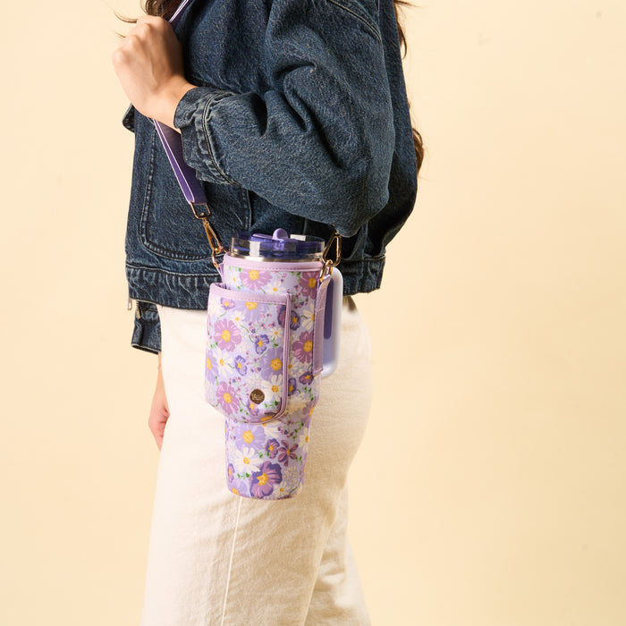 A person in a denim jacket and white pants carries the-darling-effect’s Bouquet Beauty Purple Tumbler Sling, featuring an adjustable strap for hands-free use, with a large floral water bottle. The background is plain light beige.
