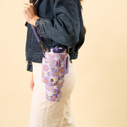 A person in a denim jacket and white pants carries the-darling-effect’s Bouquet Beauty Purple Tumbler Sling, featuring an adjustable strap for hands-free use, with a large floral water bottle. The background is plain light beige.