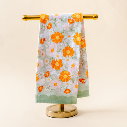 The-darling-effect's Bouquet Beauty Sage Tea Towel features orange, yellow, and white floral patterns on pale green cotton, displayed on a gold towel stand with a beige background—ideal as a decorative kitchen or tea towel.