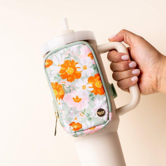 A hand holds a large white tumbler with a handle and straw, featuring the-darling-effect's Bouquet Beauty Sage Tumbler Fanny Pack—a water-resistant zippered pouch with an orange, yellow, and white floral pattern for hands-free convenience.