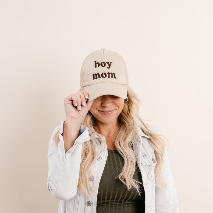 A blonde woman smiles as she adjusts The Darling Effect's Boy Mom Trucker Hat, which features trendy phrases in bold brown embroidery.