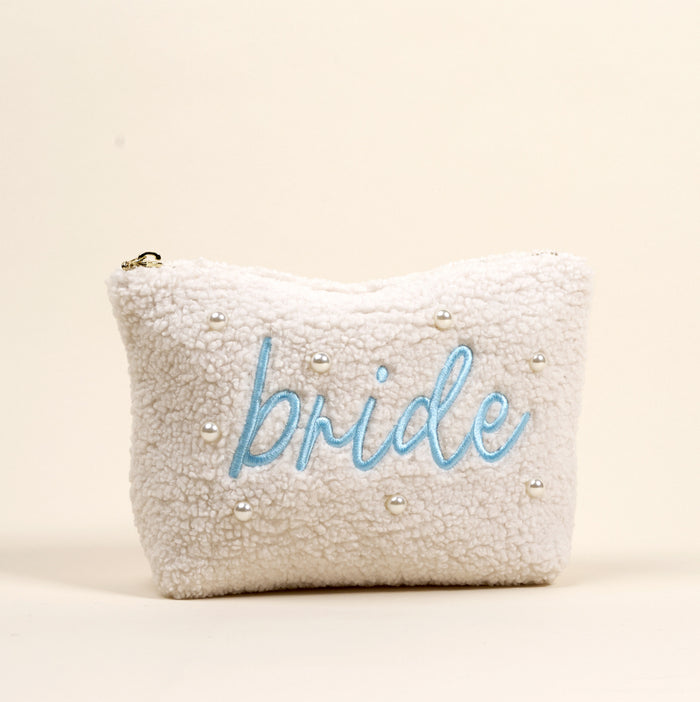 The Bride Teddy Pouch by the-darling-effect is a white sherpa pouch featuring small pearls and "bride" embroidered in light blue cursive on the front, making it a chic accessory for any bridal occasion.