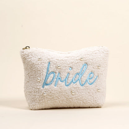 The Bride Teddy Pouch by the-darling-effect is a white sherpa pouch featuring small pearls and "bride" embroidered in light blue cursive on the front, making it a chic accessory for any bridal occasion.