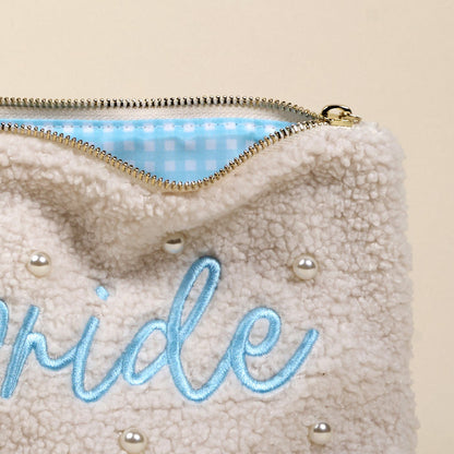 The Bride Teddy Pouch by the-darling-effect is a cream sherpa pouch with pearl accents, light blue embroidered text, a gold zipper, and a blue-and-white checkered lining—a chic accessory for any occasion.