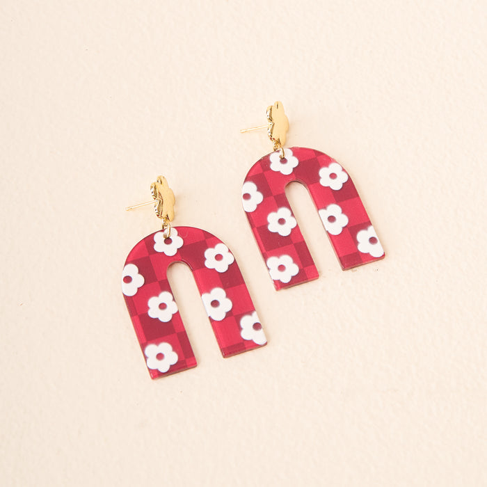 The Darling Effect's Burgundy Game Day Acrylic Earrings feature arch-shaped burgundy acrylic with white flowers and gold hooks, displayed on a light beige background.
