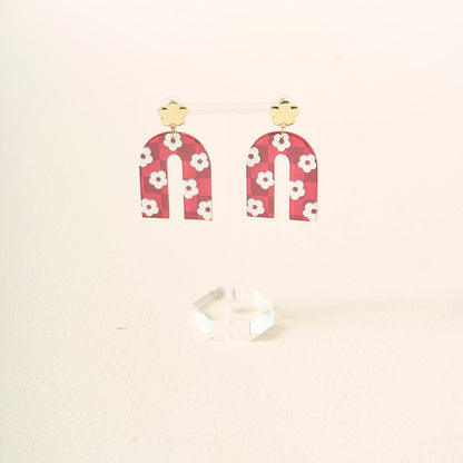 A pair of burgundy, arch-shaped acrylic earrings with white flowers and gold stud tops by The Darling Effect, displayed on a clear acrylic stand—perfect for showing off your school colors.