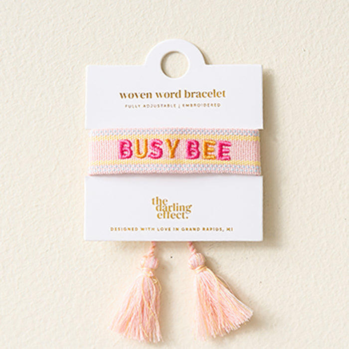 The Darling Effect’s Busy Bee Woven Bracelet features pink and yellow stripes, embroidered "BUSY BEE" text, adjustable fit, tassels, and comes on a display card. Part of The Darling Effect embroidered bracelets collection.