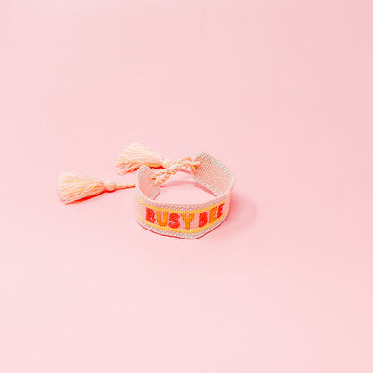 The Darling Effect's Busy Bee Woven Bracelet, shown on a pink backdrop, features "BUSY BEE" embroidered in red and yellow with tassel ends—an adorable, adjustable addition to any bracelet collection.