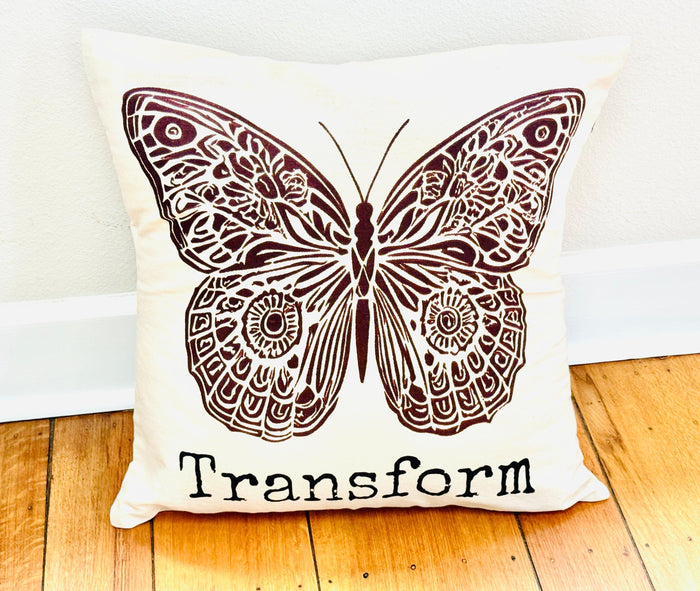Decorative  20" x 20" natural color cotton canvas pillow with a boho-style brown butterfly design and the word Transform. Handmade by Survivors.