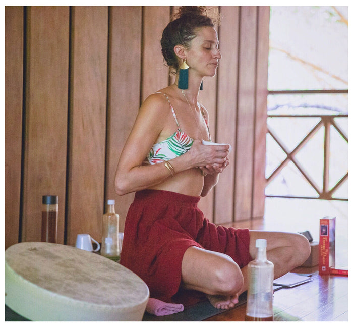 Woman sitting on a mat with a cup, surrounded by yoga props and books in a wooden room.