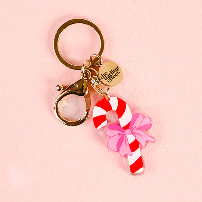 The Darling Effect’s Candy Cane Acrylic Keychain features a gold ring, lobster clasp, round “the things she did” tag, and a red-and-white candy cane charm with a pink bow on a light pink background.
