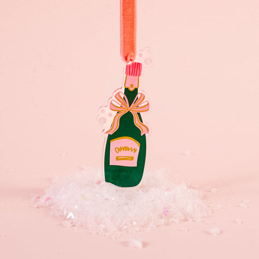The Darling Effect's Champagne Bottle Holiday Tree Ornament features a festive design with a pink bow, hanging above faux snow—perfect for holiday décor and celebrations, all set against a light pink background.