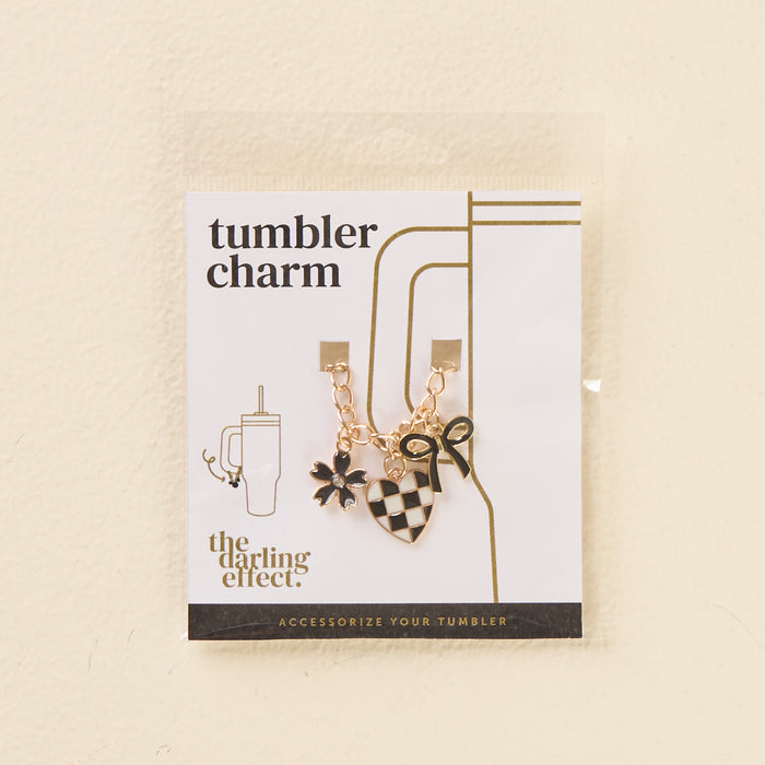 The Checkmate Tumbler Charm by the-darling-effect features a gold chain with three charms—a black flower, a black-and-white checkered heart with a bow, and a small star—perfect for personalizing your tumbler.