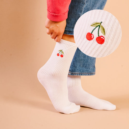 A close-up shows a person adjusting The Darling Effect Cherries Embroidered Crew Socks—white women’s socks with a small red cherry detail—while standing on a light beige background.