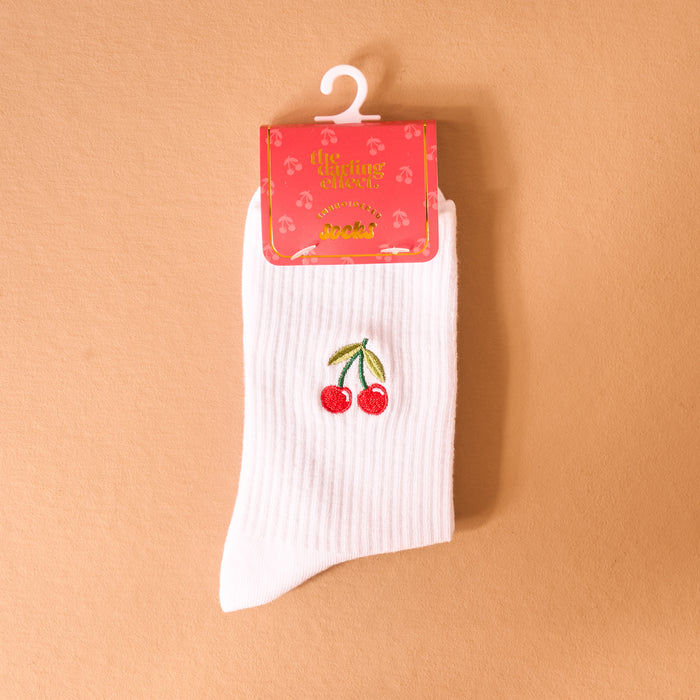 The Darling Effect's Cherries Embroidered Crew Socks, featuring a white ribbed design with cherry embroidery, are shown on a light brown background. The women's socks come packaged with a pink label decorated with yellow text and cherry illustrations.