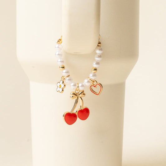 The Cherry Tumbler Charm by the-darling-effect is a beaded bracelet with gold accents, pearl-like beads, daisy and red heart charms, plus a gold cherry charm—perfect as tumbler accessories and shown draped over a white cup.